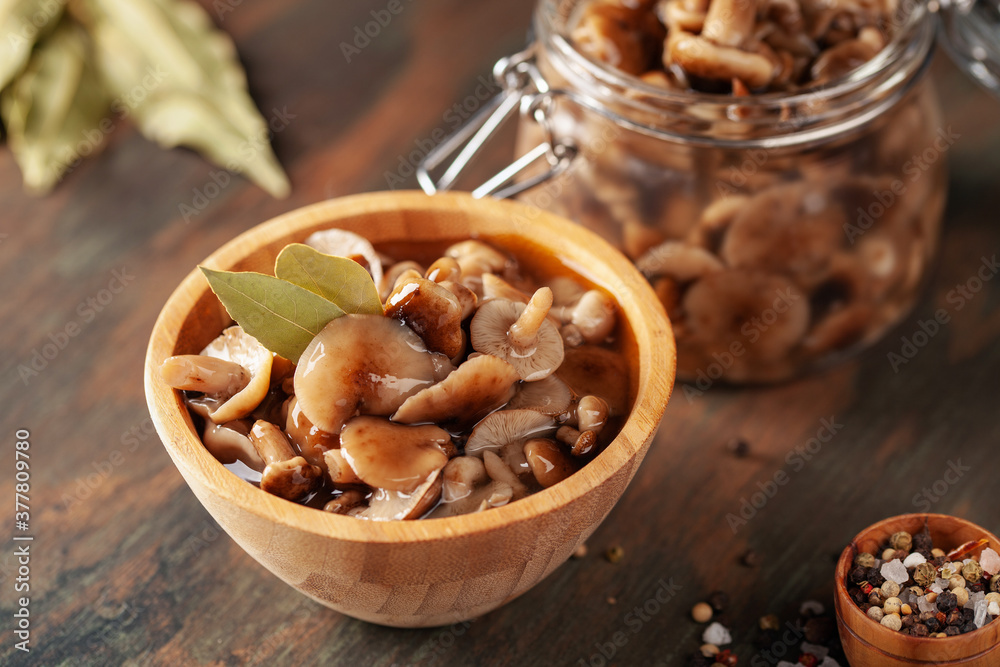 Homemade pickled honey agarics in wooden bowl and open glass jar, spices, spoon, napkin, on wooden table. Marinated mushrooms are valuable source of protein. Fermented healthy food. Homemade pickled honey agarics in wooden bowl and open glass jar, spices, spoon, napkin, on wooden table. Marinated mushrooms are valuable source of protein. Fermented healthy food.