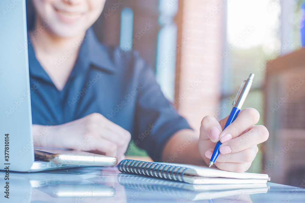 Woman is writing on a notebook with a pen and she is using a mobile phone in the office.