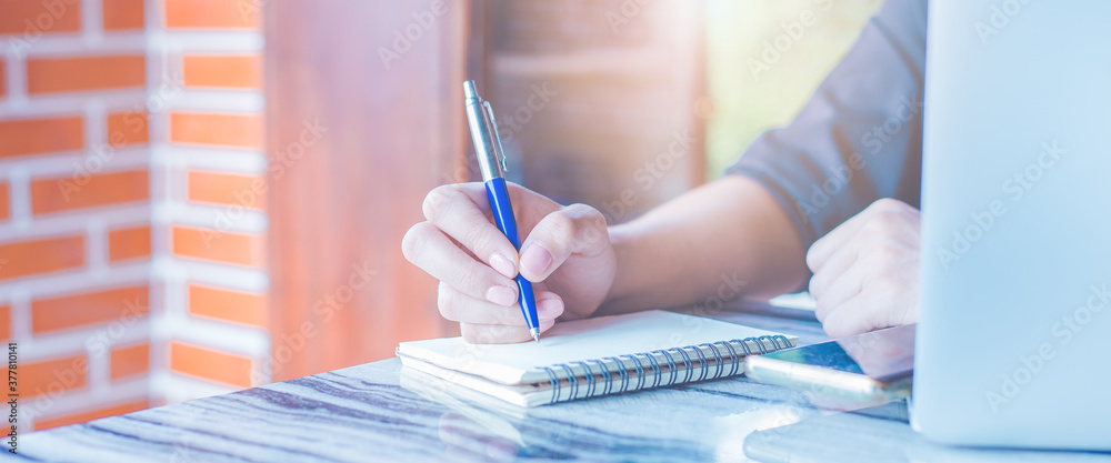 Woman is writing on a notebook with a pen and she is using a mobile phone in the office.