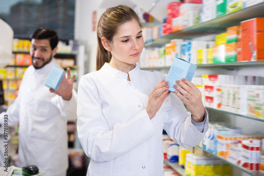 Positive female specialist is attentively looking medicines near shelves in pharmacy