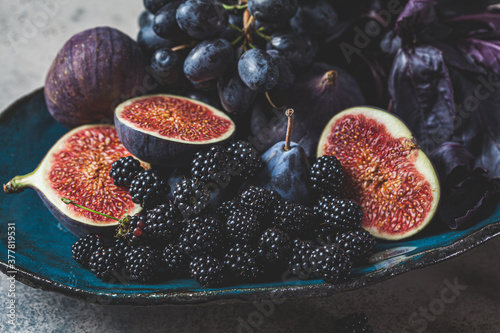 Fototapeta Naklejka Na Ścianę i Meble -  Purple fruits and berries in blue dish, gray background. Purple food background. Figs, grapes, plums and blackberries in a ceramic plate.