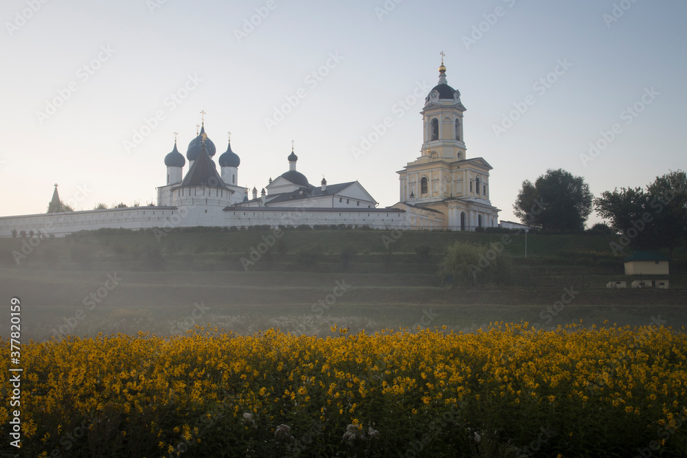 A Russian Orthodox monastery on misty sunrise. A meadow with yellow flowers in the foreground. Clear colourful sky. No people