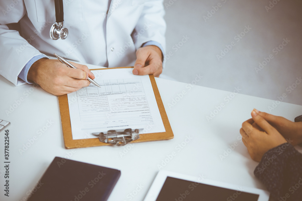 Unknown red-bearded doctor and patient woman discussing current health examination while sitting in sunny clinic, close-up. Medicine concept