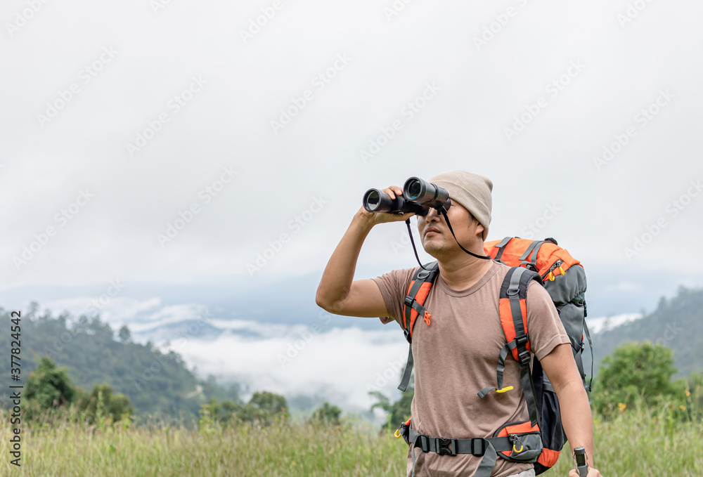 Young man with backpack and holding a binoculars looking on top of mountain with foggy landscape