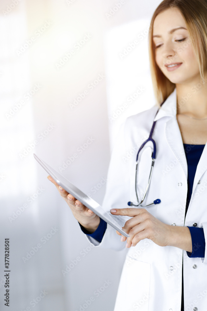 Young woman-doctor is holding a tablet computer in her hands, while standing in a sunny clinic. Portrait of friendly female physician with a stethoscope at work. Perfect medical service in a hospital Young woman-doctor is holding a tablet computer in her hands, while standing in a sunny clinic. Portrait of friendly female physician with a stethoscope at work. Perfect medical service in a hospital