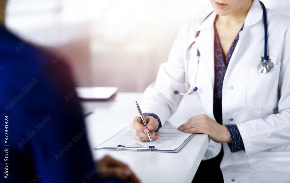 Unknown woman-doctor is writing some medical recommendations to her patient, while they are sitting together at the desk in the sunny cabinet in a clinic. Physician is using a clipboard, close-up Unknown woman-doctor is writing some medical recommendations to her patient, while they are sitting together at the desk in the sunny cabinet in a clinic. Physician is using a clipboard, close-up