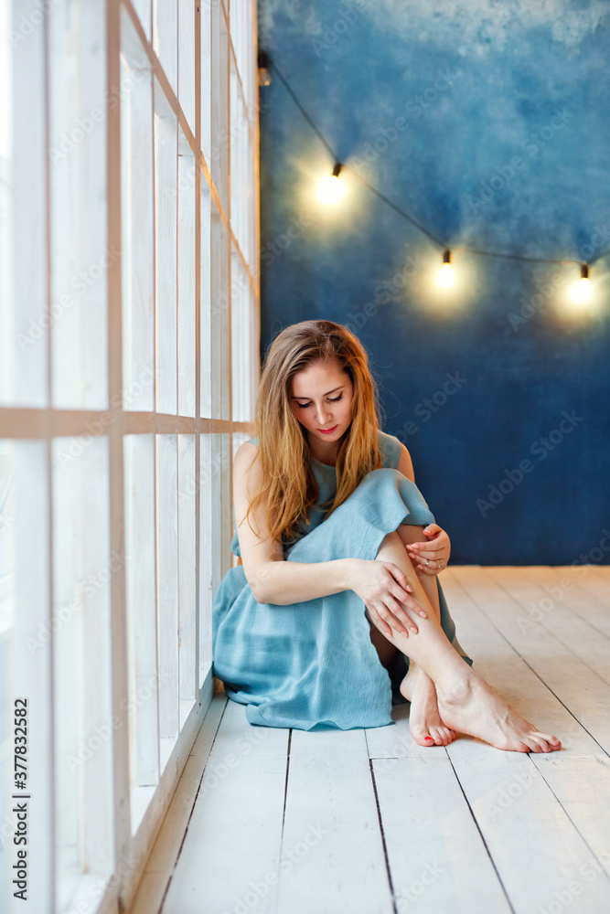 Fototapeta premium A beautiful young woman at the window, sitting on the floor with a sad face, looking away in frustration. Depression