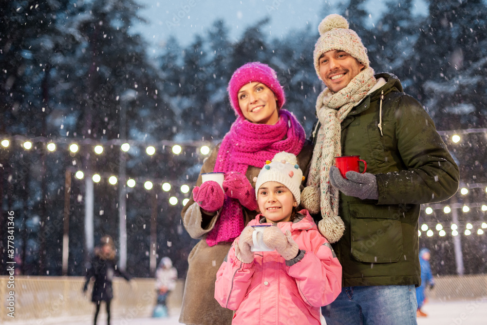 Obraz premium christmas, family and leisure concept - happy mother, father and daughter drinking hot tea at outdoor skating rink in winter