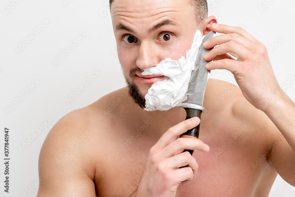 Young caucasian man shaving beard with knife on white background