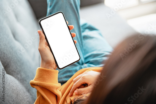 Woman using mobile smartphone with blank white screen on a sofa in living room, copy space. photo