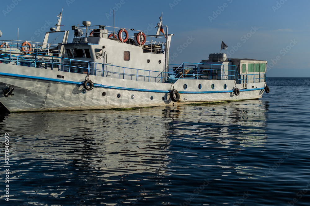 white vessel ship boat with blue stripes on pier in bay of lake baikal in the light of the sun with reflections in water