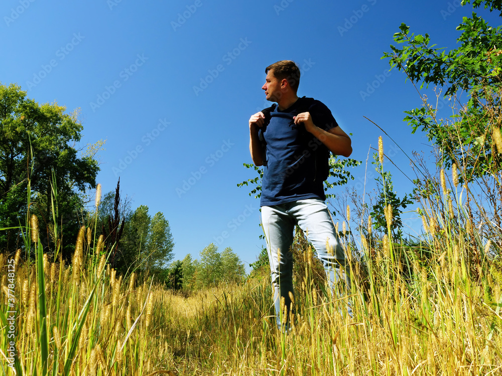 A man tourist with a backpack looks around while standing on a path in summer nature. A man tourist with a backpack looks around while standing on a path in summer nature.