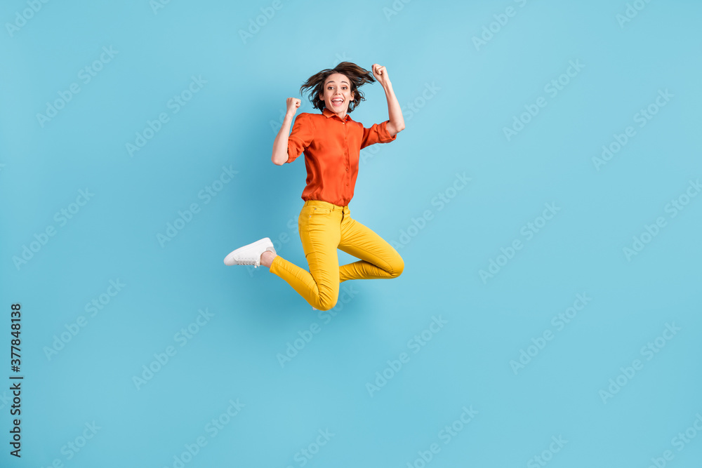 Full body photo of attractive funny crazy lady jumping high up fists hands rejoicing best win ecstatic emotions wear orange shirt yellow trousers sneakers isolated blue color background Full body photo of attractive funny crazy lady jumping high up fists hands rejoicing best win ecstatic emotions wear orange shirt yellow trousers sneakers isolated blue color background