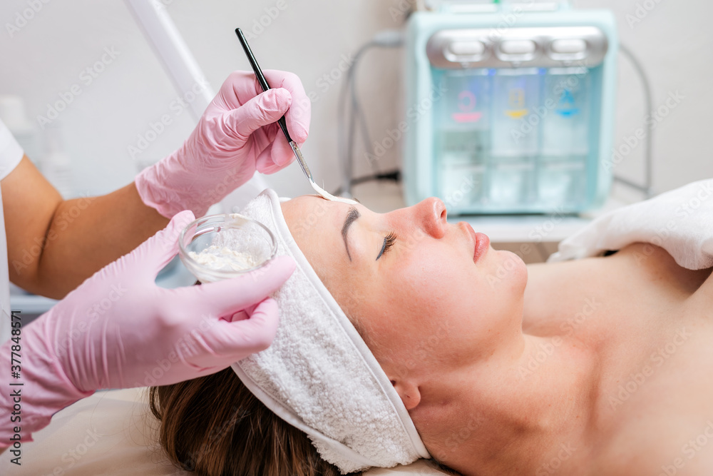 The cosmetologist in medical gloves holds a bowl with a mask, applying cosmetics with a brush on the client's face. Side view. Concept of professional cosmetology and treatment The cosmetologist in medical gloves holds a bowl with a mask, applying cosmetics with a brush on the client's face. Side view. Concept of professional cosmetology and treatment