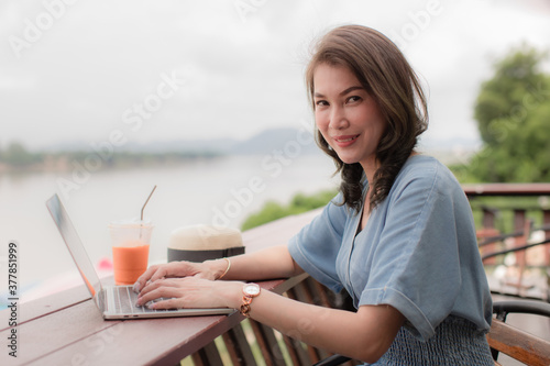 Middle age beautiful Asian woman sitting and drinking coffee in coffee shop beside river and mountain view in background while using a laptop notebook computer to work. Idea for freelancer lifestyle