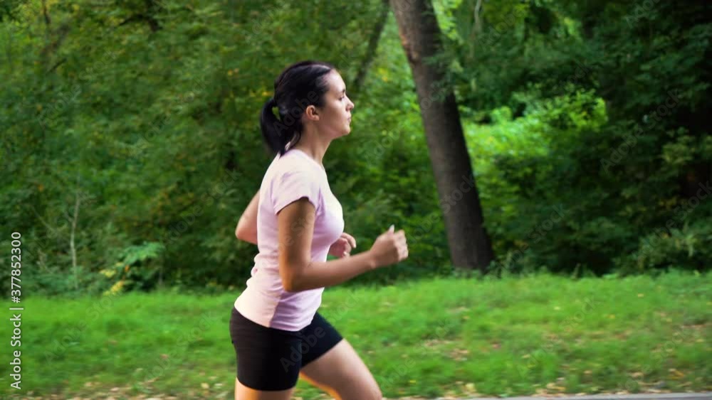 Young woman in sportswear jogging in summer park, green trees on background. Dolly shot active female training in the morning. Concept of sport