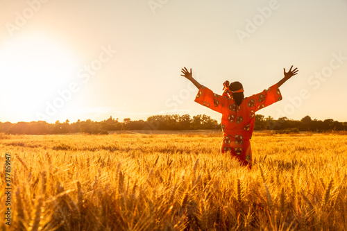 African woman in traditional clothes standing arms raised in a field of crops at sunset or sunrise