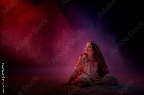 Small girl in white dress in dark studo during photoshoot with flour. Young model posing with white dust and black background. Clouds of white fog with colour light.