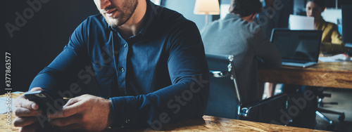 Business office concept. Portrait of handsome manager working at the table with mobile phone. Three employees at background. Wide screen, panoramic