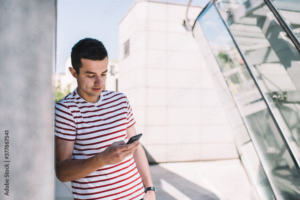 Stylish young man browsing mobile phone while standing near contemporary building in street