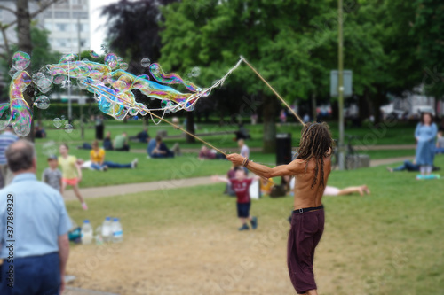 Seifenblasenkünstler in einem Park voller Menschen. Mann mit langen Dreadlocks und nacktem Oberkörper kreiert große, bunte, transparente Seifenblasen. Zuschauer freuen sich. Straßenkünstler in action.