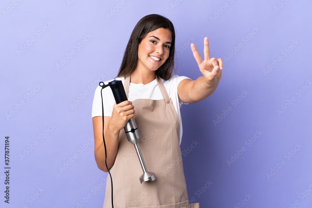 Brazilian woman using hand blender isolated on purple background smiling and showing victory sign Brazilian woman using hand blender isolated on purple background smiling and showing victory sign