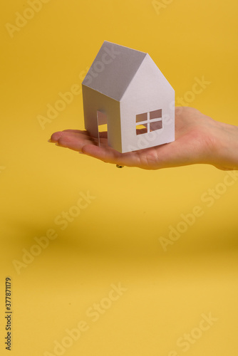 White family paper house in man hand on yellow background paper. Minimalistic style. Copy space. View from above. Vertical orientation.