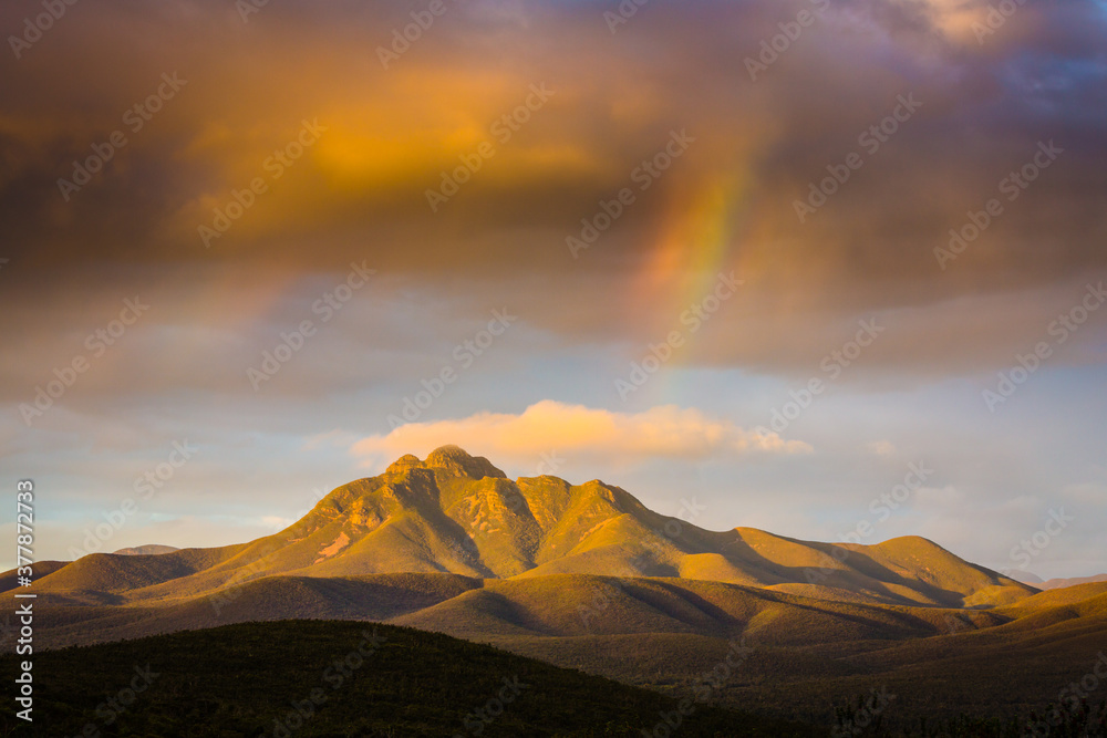 Landscape showing mountains in the the Stirling Range in West Australia at sunset. The clouds have an orange colour in the setting sun and a small rainbow is visble.