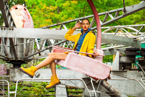 Fashion model in a bright yellow coat sits on a carousel in an amusement park