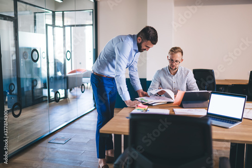 Professional male executive managers collaborating on accounting papers communicate near table desktop with mockup laptop computer at front, smart casual employees discussing financial report