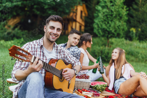 Students life.Four young attractive person chiiling in the park and one of them play at guitar and smilling