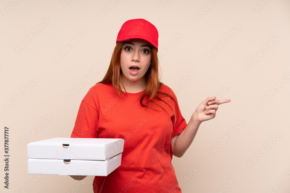Pizza delivery teenager girl holding a pizza over isolated background surprised and pointing finger to the side Pizza delivery teenager girl holding a pizza over isolated background surprised and pointing finger to the side