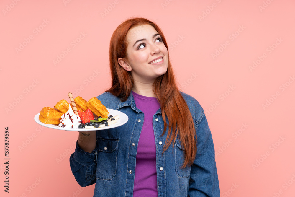 Redhead teenager girl holding waffles over isolated pink background looking up while smiling