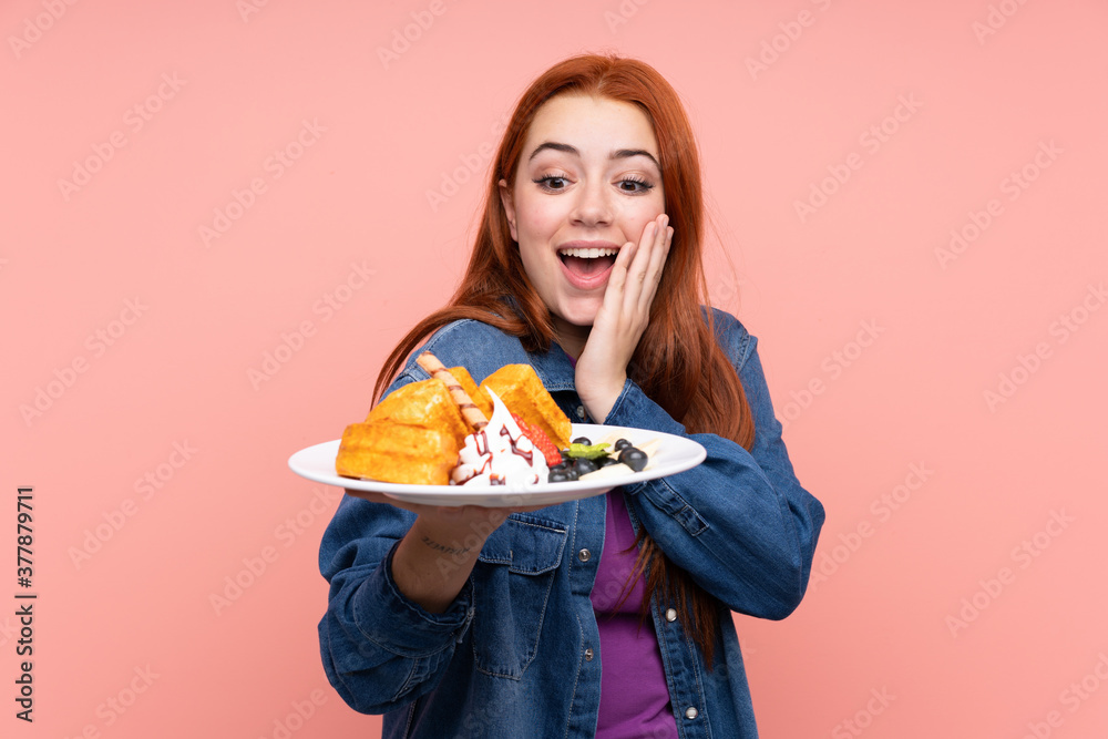 Redhead teenager girl holding waffles over isolated pink background with surprise and shocked facial expression