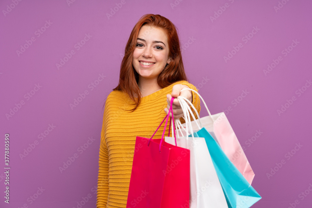 Redhead teenager girl over isolated purple background holding shopping bags and giving them to someone