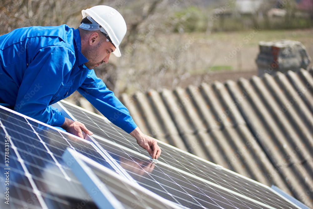 Male worker in blue suit and protective helmet installing stand-alone solar photovoltaic panel system. Electrician mounting solar module on roof of modern house. Alternative energy ecological concept. Male worker in blue suit and protective helmet installing stand-alone solar photovoltaic panel system. Electrician mounting solar module on roof of modern house. Alternative energy ecological concept.
