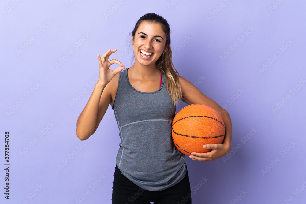 Young hispanic woman over isolated purple background playing basketball and making OK sign
