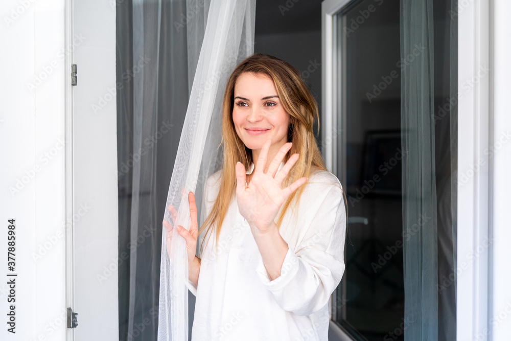 Woman at home looking through window and waving to somebody Woman at home looking through window and waving to somebody