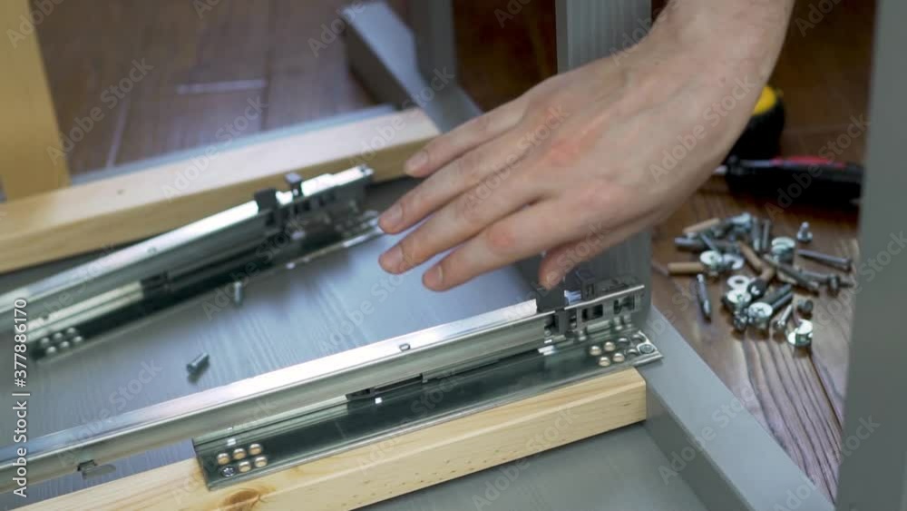 Carpenter Furniture Assembler Checks The Work of A Metal Sliding Structure For a Drawer of a Prefabricated Cabinet, Close-up. Assembly of Furniture. Carpenter Furniture Assembler Checks The Work of A Metal Sliding Structure For a Drawer of a Prefabricated Cabinet, Close-up. Assembly of Furniture.