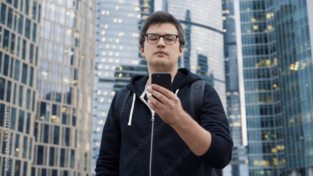 Man in eyeglasses holding a phone on background of business centre. Man dressed in casual clothes texting in the phone wearing a black hoodie Man in eyeglasses holding a phone on background of business centre. Man dressed in casual clothes texting in the phone wearing a black hoodie