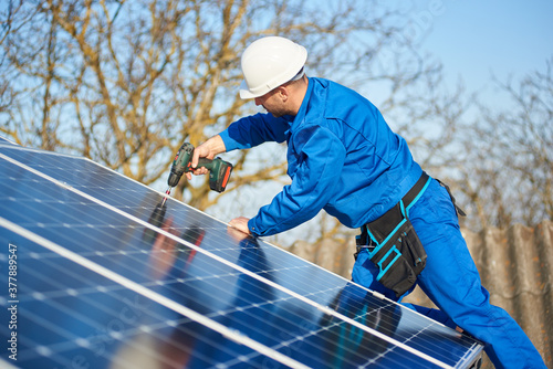 Male engineer in blue suit and protective helmet installing solar photovoltaic panel system using screwdriver. Professional electrician mounting blue solar module on roof of modern house.