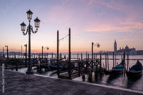 A lamp on the Riva degli Schiavoni waterfront with docked gondolas and San Giorgio Maggiore basilica in the background during a sunsrise in Venice, Italy