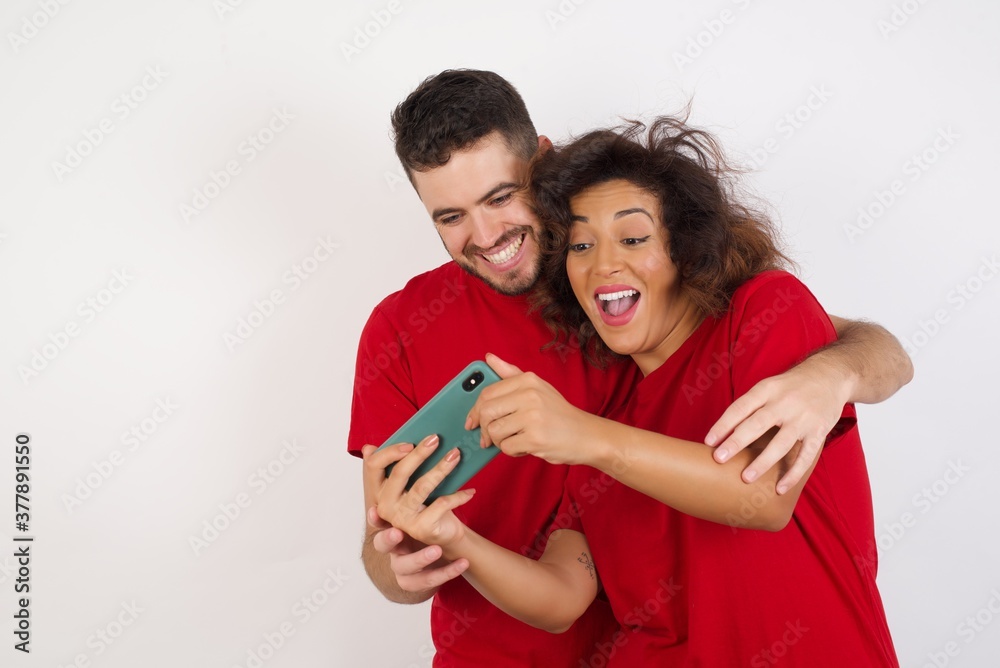 Young beautiful couple wearing red t-shirt on white background holding in hands cell playing video games or chatting Young beautiful couple wearing red t-shirt on white background holding in hands cell playing video games or chatting