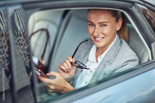 Portrait of a happy mature business woman sitting on back seat in the car and using smartphone, she is looking at camera and smiling
