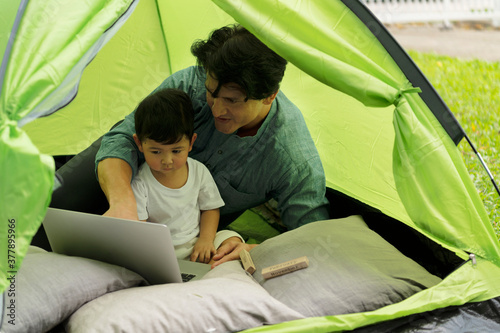 Joyful young father in casual wear sets up a camping tent in family home outdoor garden with happy little son and having fun together with children toy and laptop