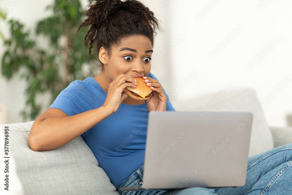 African Girl Eating Burger Using Laptop Sitting On Couch Indoor African Girl Eating Burger Using Laptop Sitting On Couch Indoor