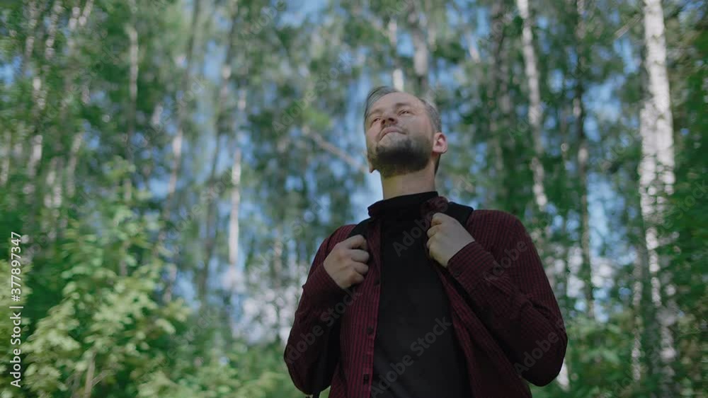 Young handsome bearded man with a backpack walking along a forest path looking around with a smile. The sun's rays break through the leaves of the trees. Low angle high quality 4k footage Young handsome bearded man with a backpack walking along a forest path looking around with a smile. The sun's rays break through the leaves of the trees. Low angle high quality 4k footage