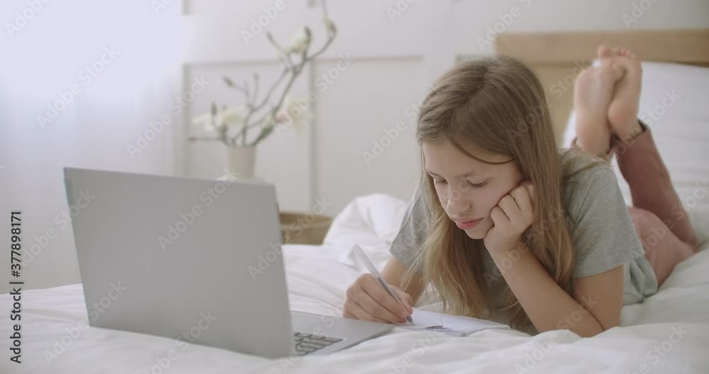 little girl is drawing and writing in copybook, lying on bed in front of laptop in her room, waiting online lesson little girl is drawing and writing in copybook, lying on bed in front of laptop in her room, waiting online lesson