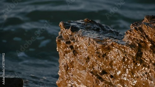 Waves breaking on the rock of the coastline. Wet brown sharp rock under the water. Slow motion, close up, shallow depth of field