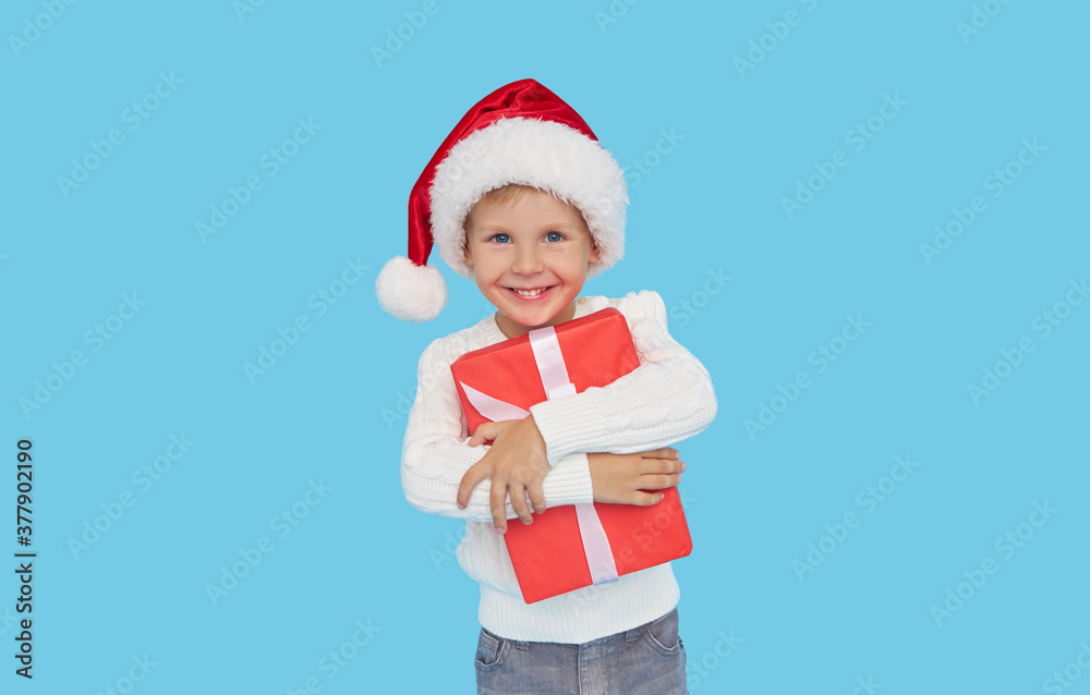 Portrait of a satisfied little child boy in christmas Santa hat. laughing isolated over blue background. Holds a gift box. Preparing for the New Year holidays Portrait of a satisfied little child boy in christmas Santa hat. laughing isolated over blue background. Holds a gift box. Preparing for the New Year holidays